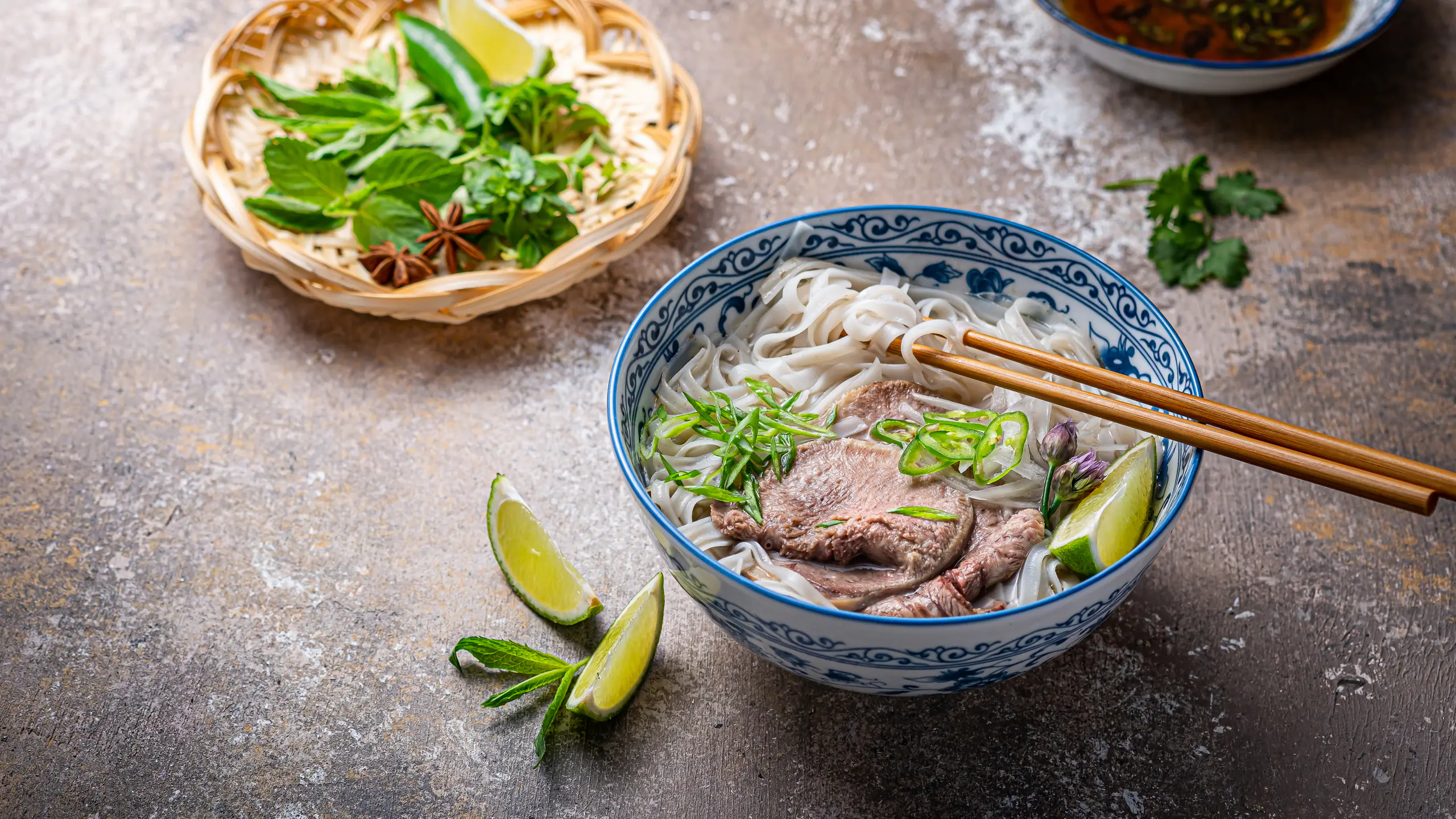 A blue-and-white bowl of phở with sliced beef, rice noodles, spring onion, chilli and lime, beside a basket of fresh herbs and star anise