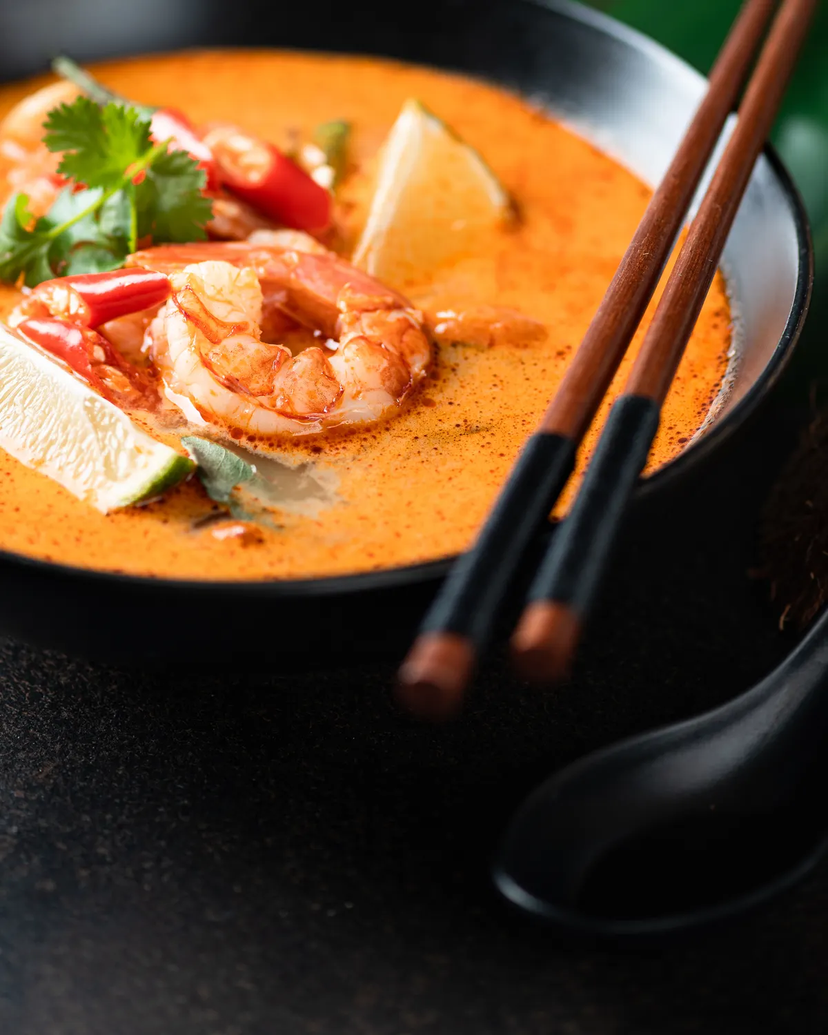 A bowl of coconut prawn soup with lime, red chilli and coriander, wooden chopsticks resting across the rim